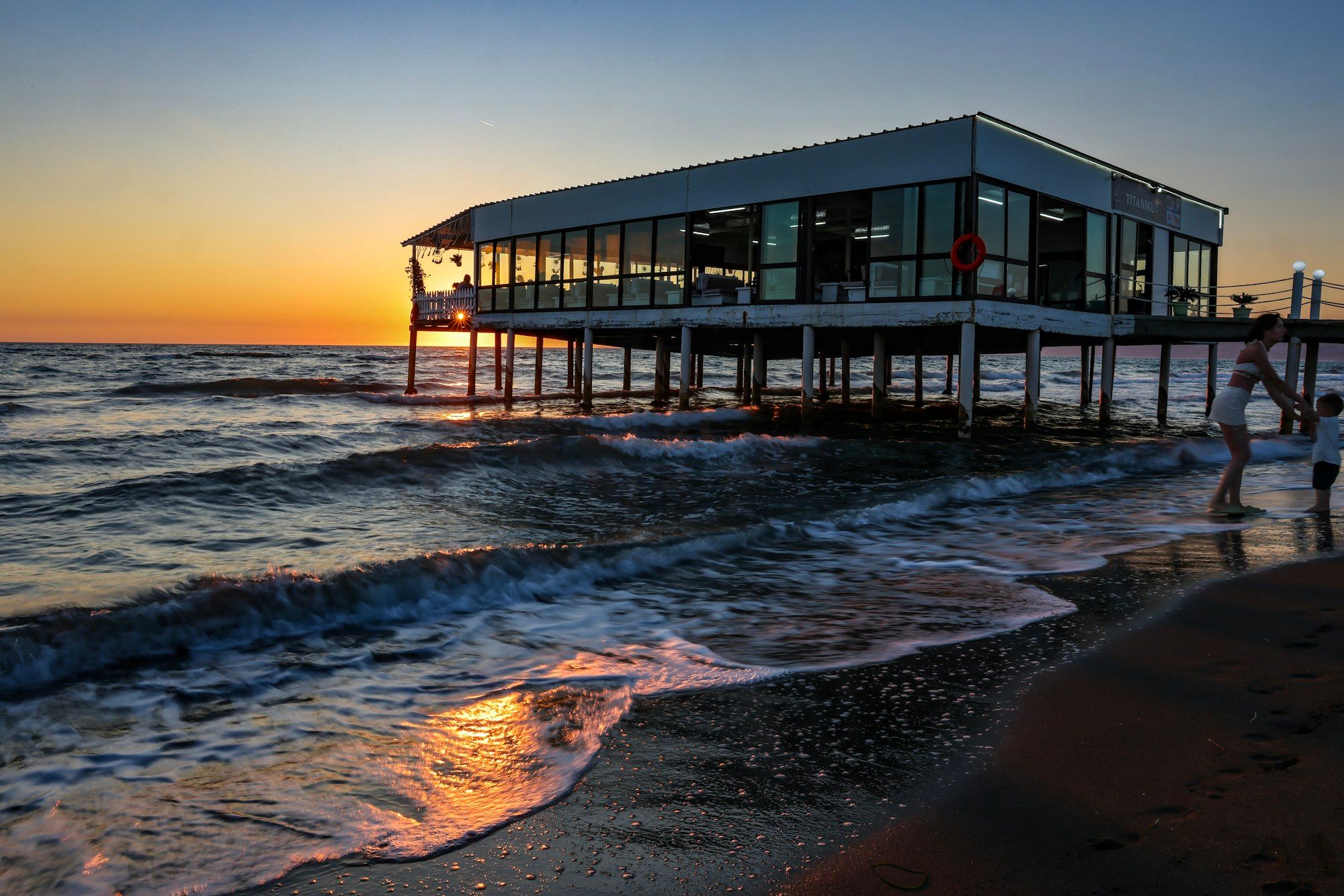 A restaurant on stilts above the sea at sunset, seen from the shore.