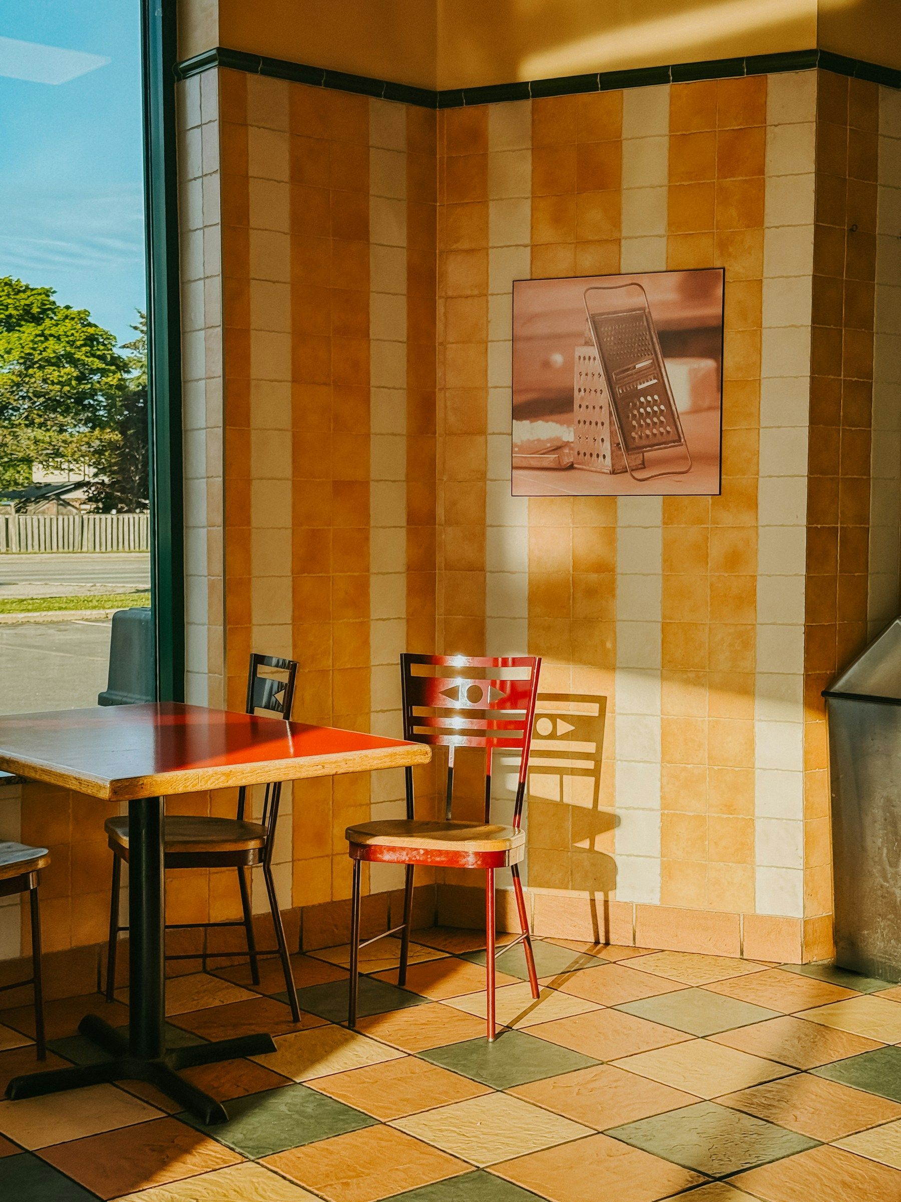 A sunlit café corner with a red table, a red chair and striped tiled walls.