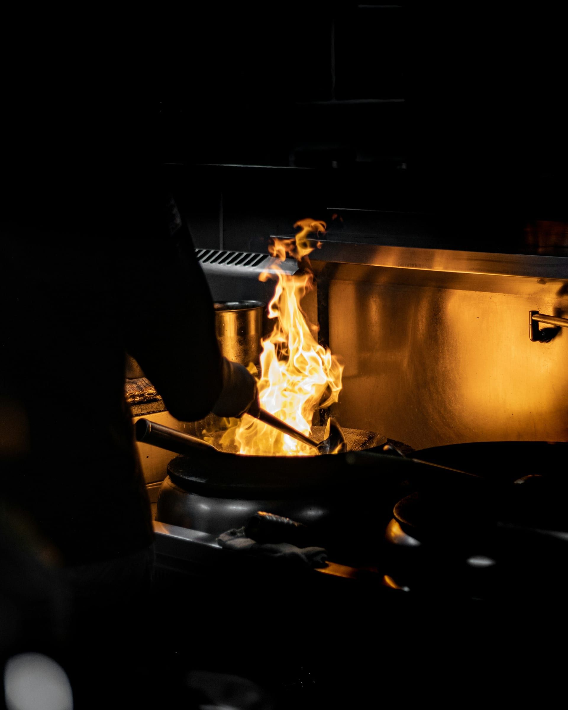 A cook tossing a wok over a tall flame in a dark kitchen.